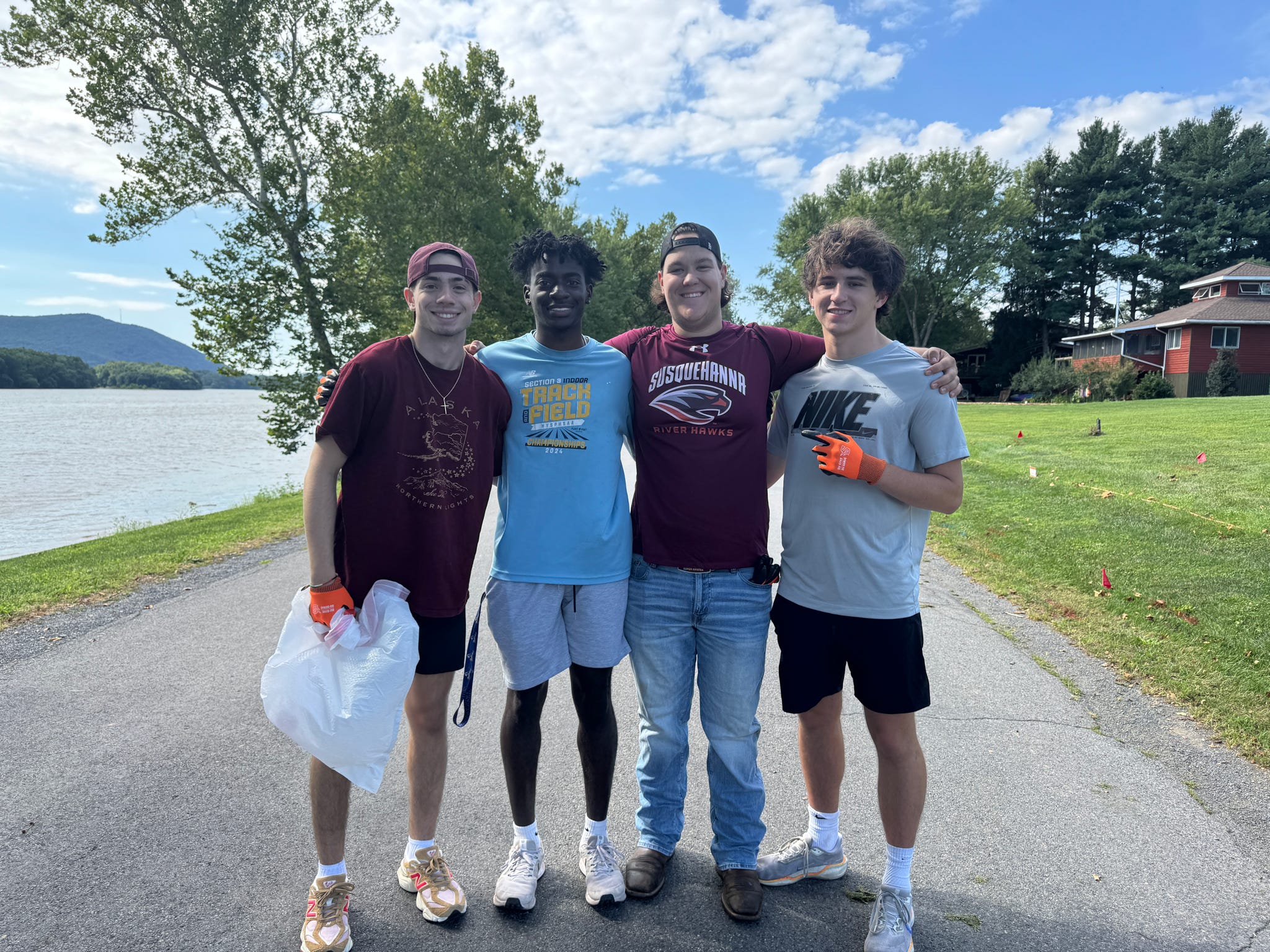 Four students standing together outdoors beside a lake, with gloves and a collection bag